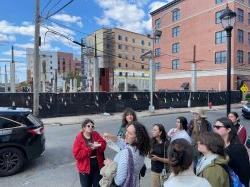 一群学生站在建筑工地前的人行道上. 学生们在奥兰治大学的徒步旅行中做笔记