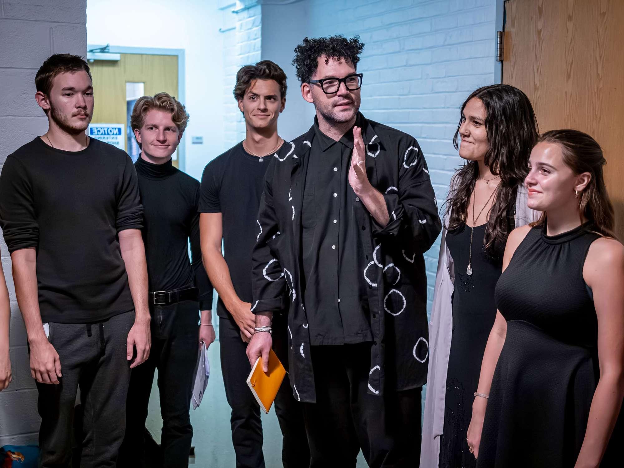 The play’s director stands in the center of five students, all dressed in black.